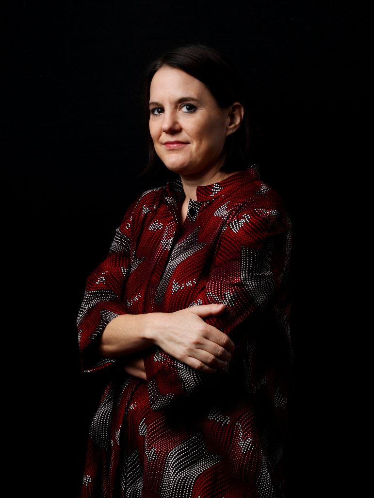A picture of a woman in a patterned red, white, and black dress in front of a black background. She has her arms crossed and is looking from the side towards the camera.